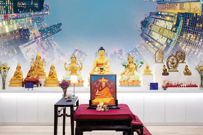 Imagem de um altar budista kadampa com o trono à frente onde se encontra um quadro com a imagem de Venerável Geshe Kelsang Gyatso Rinpoche. Ao fundo, como uma montagem, vêm-se prédios modernos como se observados do nível da rua, dando uma impressão de cidade grande.